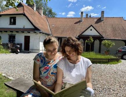 Kolorowa fotografia dwóch młodych, uśmiechniętych kobiet na tle oddziału Rydlówka. W dłoniach trzymają album.