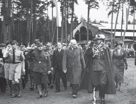 Czarno-białą fotografia. Osoby w mundurach, strojach harcerskich oraz płaszczach na tle drzew.